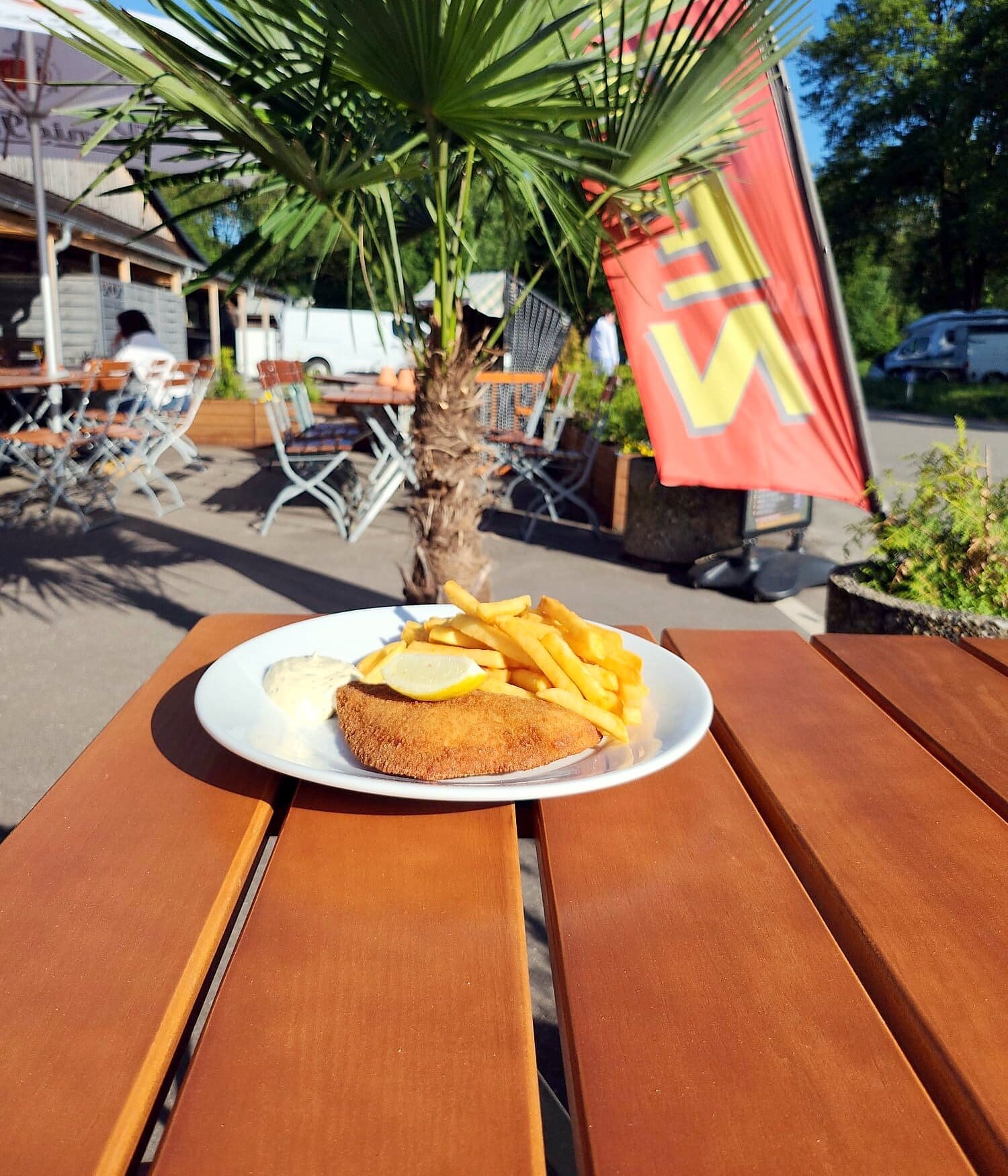 Paniertes Gericht mit Pommes und Zitrone auf einem Holztisch im Biergarten, dahinter Palme, Klappstühle und eine rote Fahne